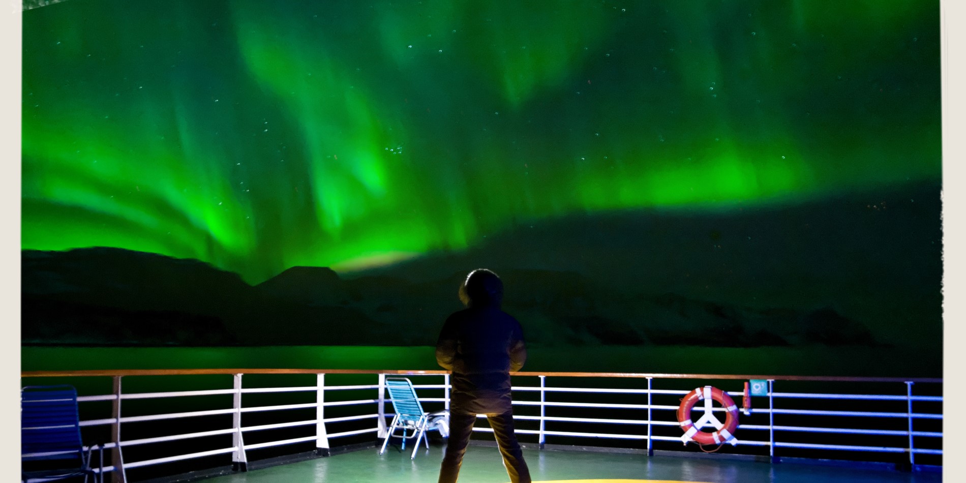 Man på däck på Hurtigrutens fartyg som trafikerar den norska kusten. Över honom är himlen färgad i grönt – norrskenet dansar