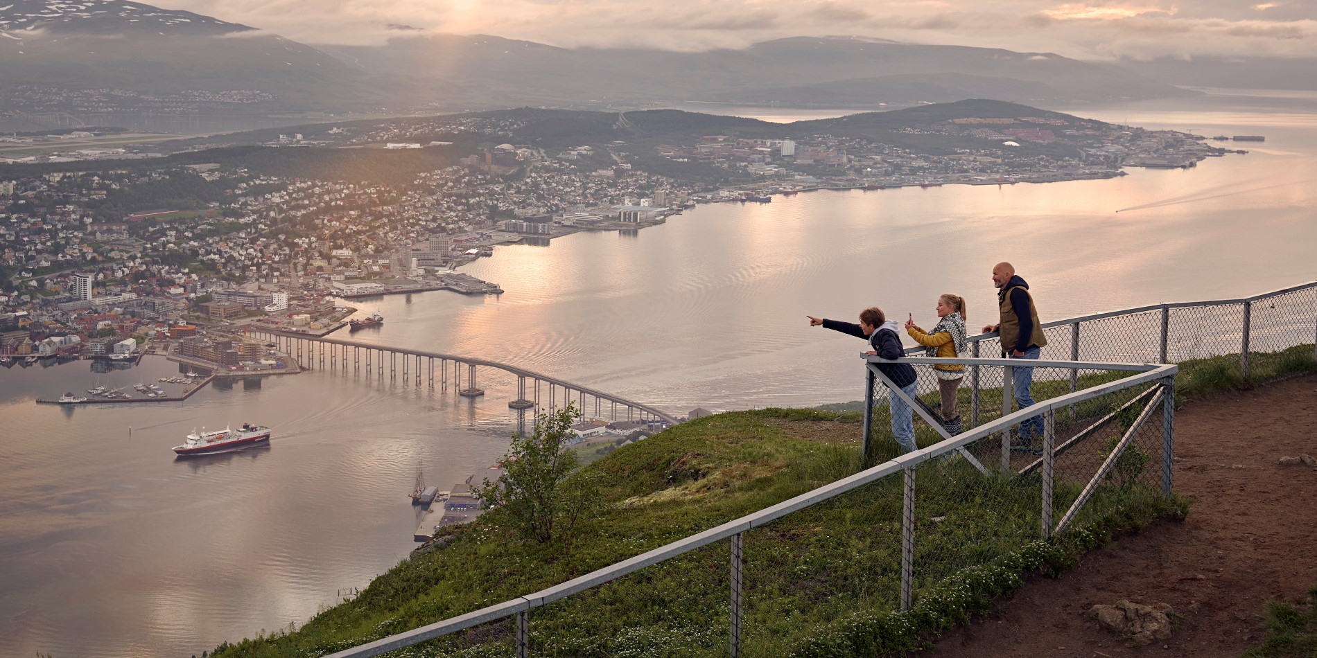 Tre barn står bakom ett staket på toppen av fjället i Tromsø. De blickar ut över Tromsø centrum.