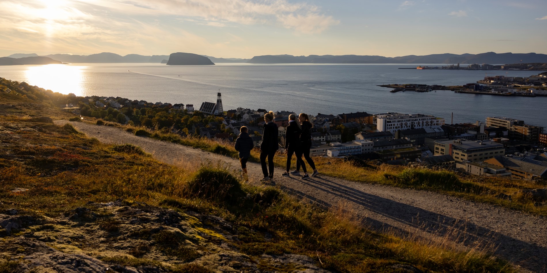 Fyra personer promenerar längs fjällsluttningen i solnedgången. Nedanför kan de blicka ut över Hammerfest